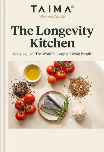 Front cover of the book 'The Longevity Kitchen' by Taima Wellness Guide, featuring a cream-colored background with dark green text and a central photograph of fresh ingredients including tomatoes, garlic, herbs, and fish.