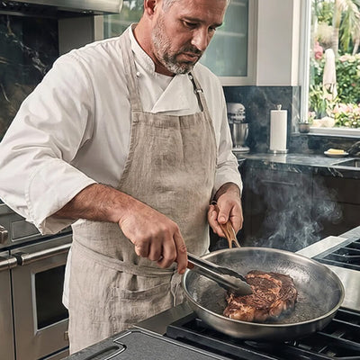 A chef in a white uniform and beige apron cooks a steak in a stainless steel pan on a gas stove in a modern kitchen.