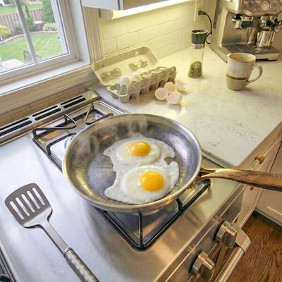 Two eggs cooking in a stainless steel skillet on a gas stovetop in a bright kitchen. A carton of eggs, cracked eggshells, a coffee maker, and a mug sit on the countertop nearby.