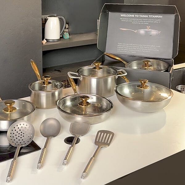 A Titanium cookware set arranged on a white countertop with its open retail box behind it. The set includes multiple pots, a wok, lids, and utensils with gold-toned handles and knobs.