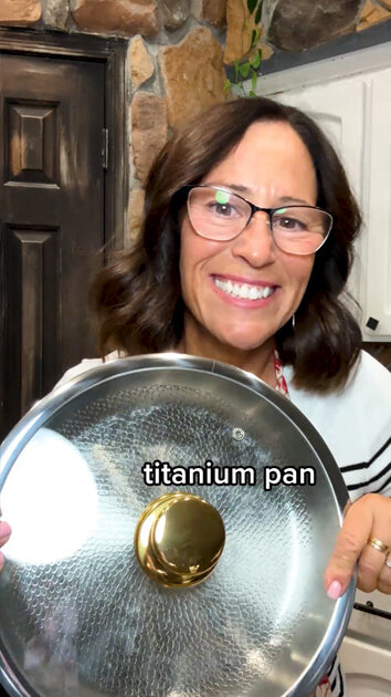 A smiling woman with glasses holds up a large hammered stainless steel pan lid with a gold knob in a kitchen setting. Text on the lid reads ' titanium pan'.