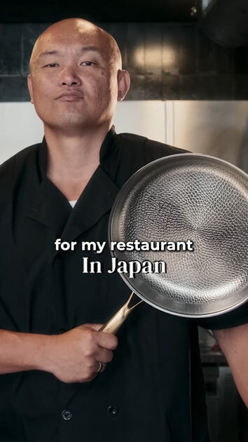 A bald male chef in a black uniform holds up a hammered stainless steel frying pan with a wooden handle. White text overlay reads 'for my restaurant In Japan'.