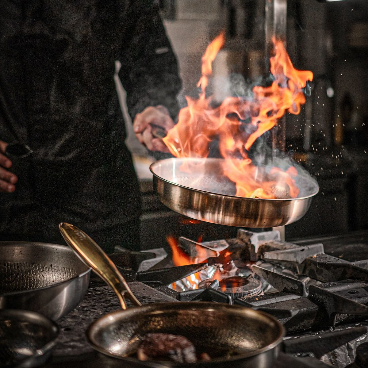 Person cooking over a flaming stove with multiple pans on a stovetop.